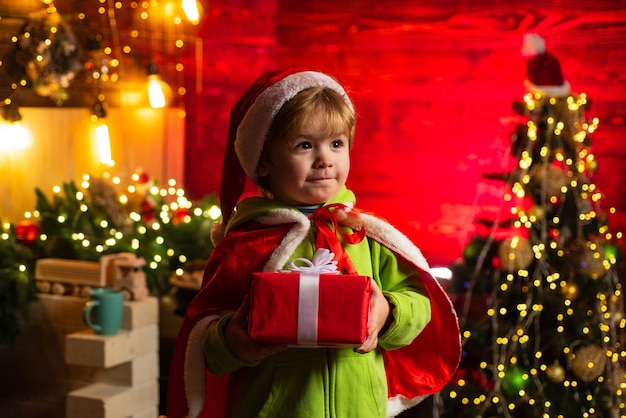 Menino feliz perto da árvore de natal com um presente de natal a criança está vestindo roupas de papai noel chr