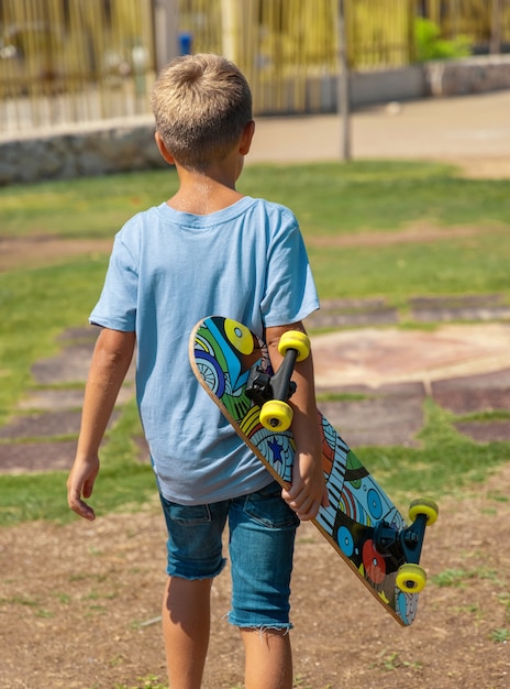 Menino feliz pré-escolar em t-shirt manter um skate nas mãos ao ar livre. linda criança fazendo esportes e se divertindo no parque. hobbies ativos, conceito de estilo de vida saudável. maquete da camiseta, vista traseira