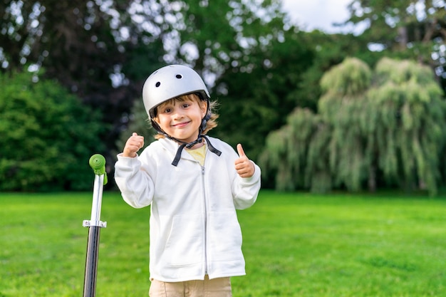 Menino feliz usando capacete de segurança, ficando perto de scooter de chute. feche o retrato de uma criança sorridente andando em um parque.