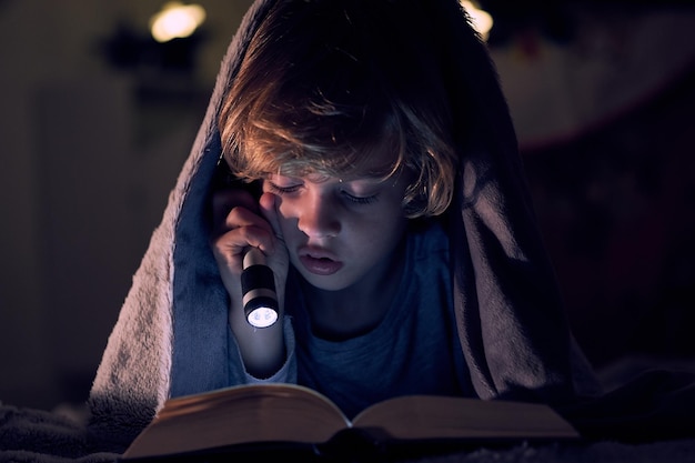 Menino focado lendo um livro com interesse e uma lanterna brilhante enquanto deitado na cama sob um cobertor quente em uma sala escura do apartamento