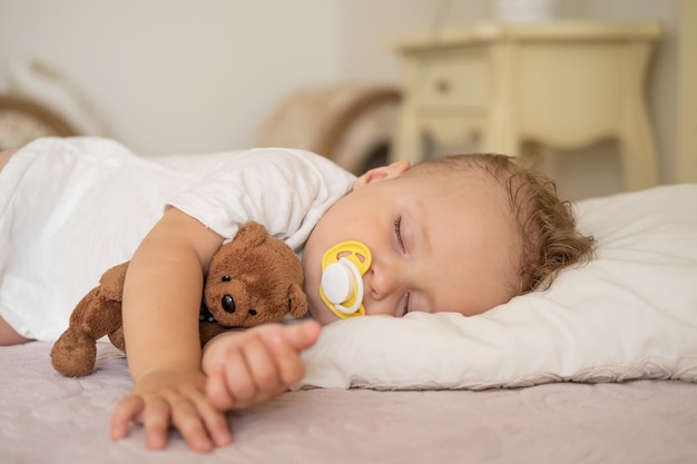 Menino fofo com mamilo dormindo na cama em casa criança abraçando o ursinho de pelúcia