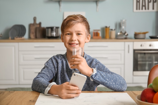 Menino fofo com um celular bebendo água na cozinha