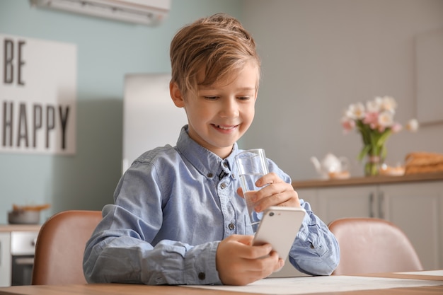 Menino fofo com um celular bebendo água na cozinha