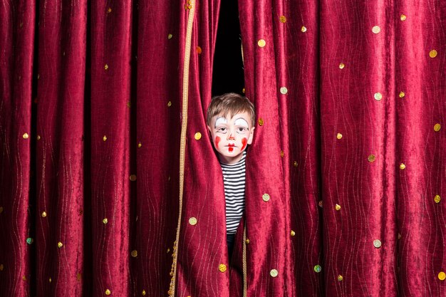 Menino fofo com uma pintura facial vermelha colorida e uma fantasia espiando por entre as cortinas do palco enquanto espera o início da pantomima