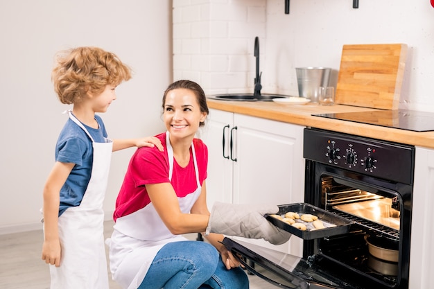 Foto menino fofo de avental conversando com a mãe dele, colocando a bandeja com biscoitos crus no forno enquanto cozinham juntos na cozinha