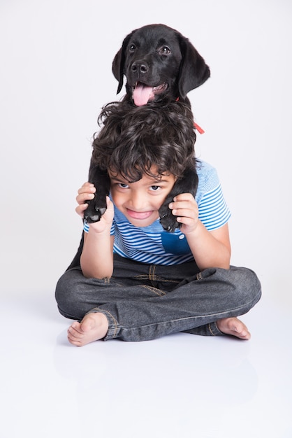 Foto menino fofo indiano ou asiático brincando com o filhote de cachorro labrador retriever preto enquanto está deitado ou sentado, isolado no fundo branco