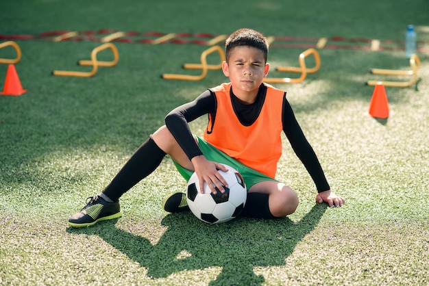 Menino hispânico fofo em uniforme de futebol com bola de futebol após um treinamento intensivo no estádio