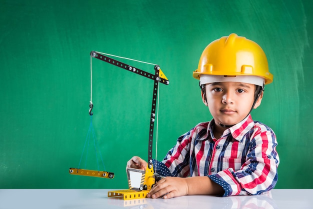 Menino indiano fofo brincando com um guindaste de brinquedo, usando um chapéu ou capacete amarelo de construção, conceito de infância e educação, isolado sobre o quadro verde