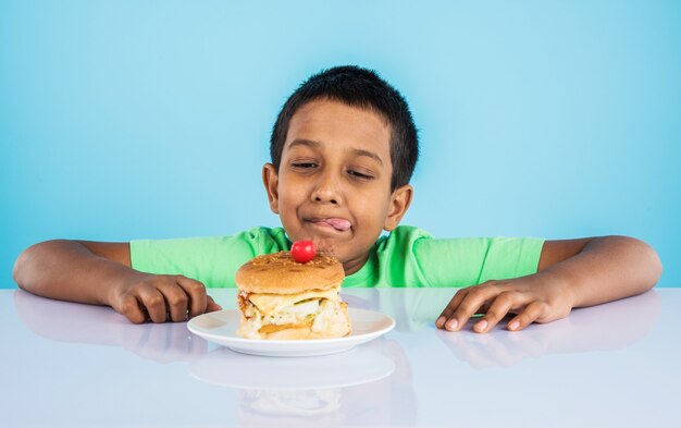 Menino indiano fofo comendo hambúrguer, pequeno menino asiático e hambúrguer, sobre fundo azul