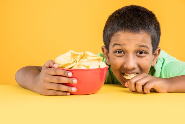 Menino indiano ou asiático bonitinho comendo batatas fritas ou bolachas de batata em uma grande tigela vermelha, sobre fundo amarelo