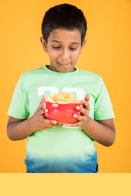 Menino indiano ou asiático bonitinho comendo batatas fritas ou bolachas de batata em uma grande tigela vermelha, sobre fundo amarelo