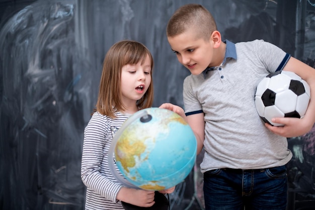 menino infantil feliz com bola de futebol e menina aprendendo sobre o mundo usando o globo da terra em pé na frente do quadro negro