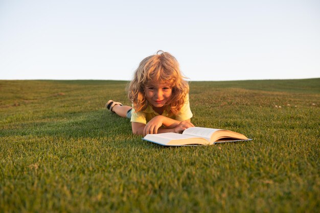 Menino inteligente lendo livro deitado na grama no fundo da grama e do céu