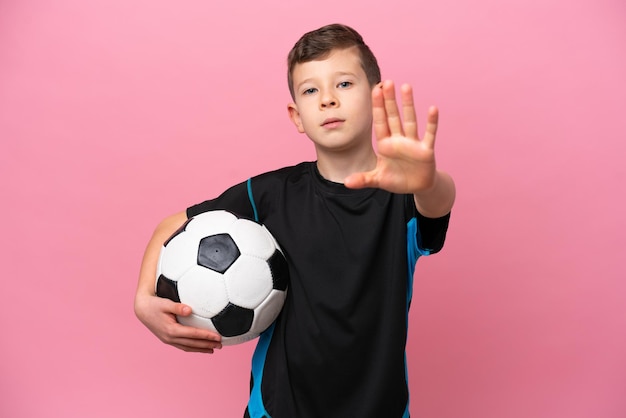 Menino jogador de futebol caucasiano isolado em fundo rosa fazendo gesto de parada