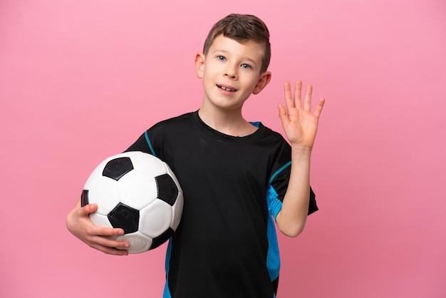 Menino jogador de futebol caucasiano isolado no fundo rosa saudando com a mão com expressão feliz