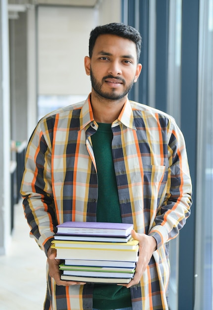 Menino jovem estudante indiano lendo livro estudando na biblioteca da faculdade com estante atrás de trabalho em atribuição ou projeto
