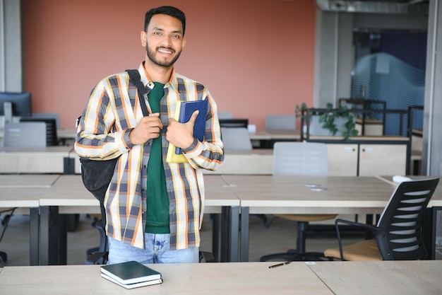 Menino jovem estudante indiano lendo livro estudando na biblioteca da faculdade com estante atrás de trabalho em atribuição ou projeto