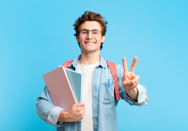 Menino jovem estudante sorrindo e parecendo feliz, despreocupado e positivo, gesticulando vitória ou paz com uma mão