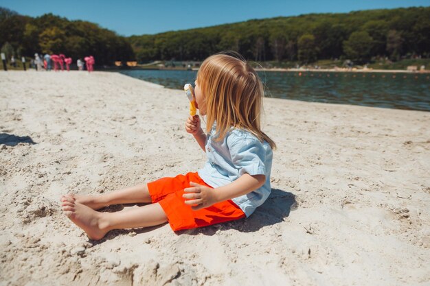 Menino loiro bonitinho com cabelo comprido comendo sorvete nas férias de verão na praia