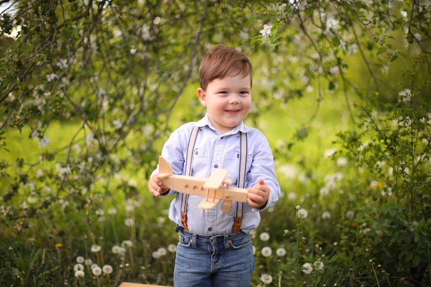 Menino loiro bonito brincando com um avião de madeira no parque de verão na grama em um dia ensolarado