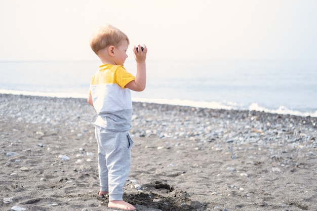 Menino loiro brincando com pedras e areia na praia em um dia ensolarado.