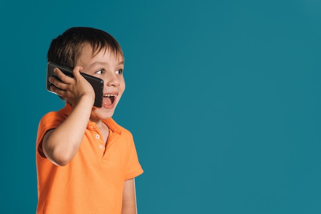 Menino loiro com uma camiseta laranja falando surpreso ao telefone sobre um fundo azul. copie o espaço.