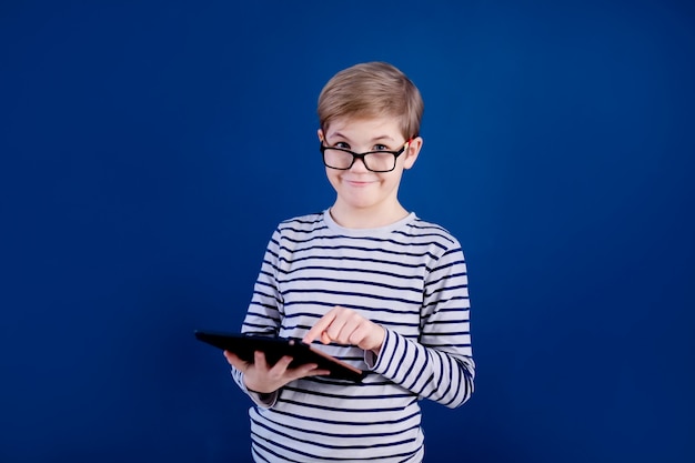 Menino loiro criança com óculos grandes, brincando com o tablet pc na parede azul. Conceito de educação.