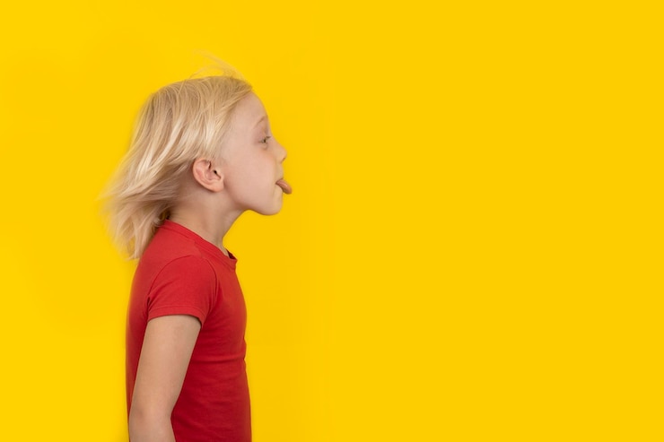 Foto menino loiro de camisa vermelha mostra a língua vista lateral do retrato em fundo amarelo copiar espaço