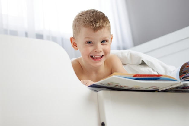 Menino loiro de olhos azuis sorri enquanto lê um livro deitado em um berço branco