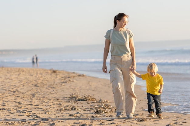 Menino loiro e sua mãe caminhando na praia à beira-mar