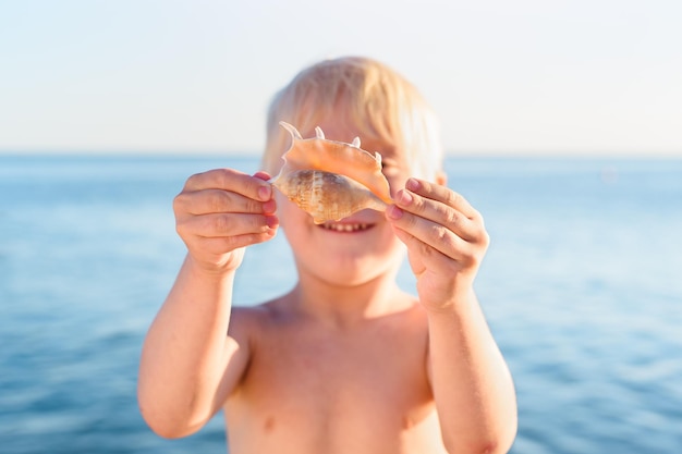 Menino loiro segurando concha e sorrindo Férias com crianças do mar Conceito de infância feliz