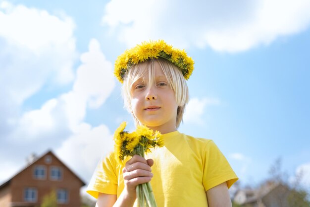 Menino louro em camiseta amarela com coroas de flores e buquê de flores contra o céu azul e casas infância feliz mundo sem guerra