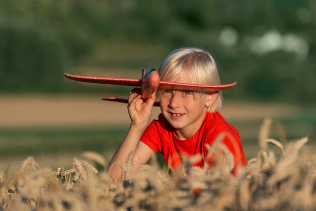 Menino louro em camiseta vermelha segura avião modelo vermelho e sentado em trigo maduro Retrato de criança com avião de brinquedo