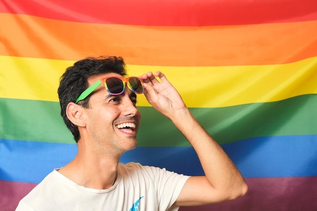 Menino moreno sorridente em camiseta branca com óculos lgbtq arco-íris, levantando os óculos no fundo da bandeira do arco-íris do orgulho gay