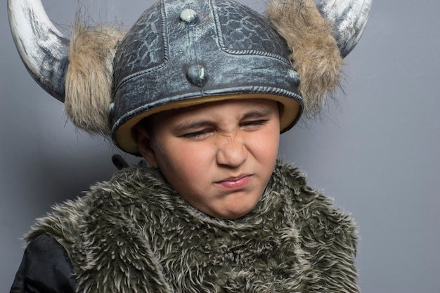 menino moreno viking em camisa de pele em idade pré-escolar em pé dentro de casa, usando chapéu de fantasia com chifres, mãos para cima, fazendo uma cara assustadora e espada