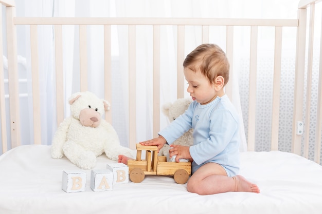 Menino no berço do quarto das crianças brincando com brinquedos de madeira em uma cama de algodão branco