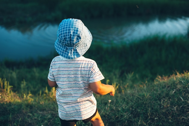 Menino olhando para um pequeno rio e jogando pedras com um chapéu azul em um campo verde