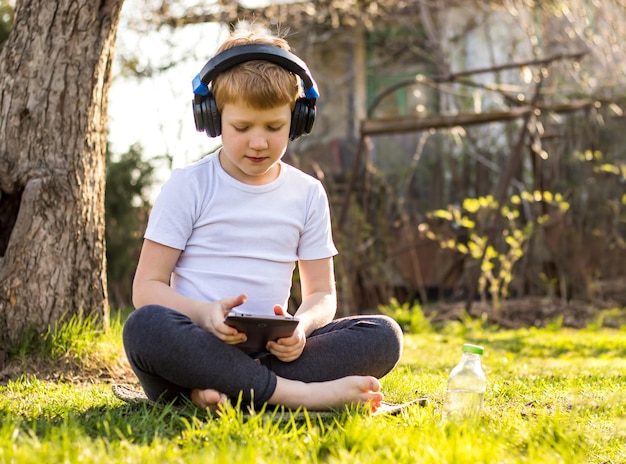 menino ouvindo música em fones de ouvido, usando dispositivo móvel, relaxando sentado na grama na natureza