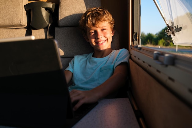 Menino positivo olhando para a câmera com um sorriso e usando um laptop enquanto sentado perto da janela do acampamento em um dia de fim de semana de verão