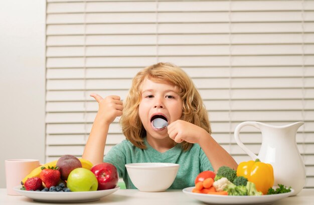 Menino pré-adolescente lamber colher na cozinha na mesa comendo legumes e frutas durante o jantar