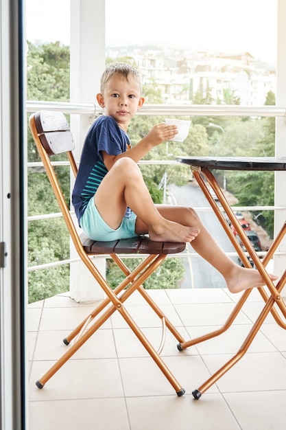 Menino pré-escolar bebe leite morno da caneca sentado na cadeira no início da manhã na varanda do hotel