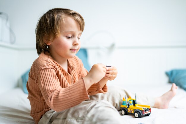 Menino pré-escolar brincando com o construtor, sentado em uma cama na sala. Criança passando algum tempo durante a quarentena. Conceito de jogos de desenvolvimento, espaço de cópia.