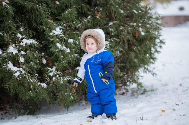 Menino rindo de um ano de idade com roupa de neve quente, caminhando no parque de inverno com uma neve branca. Primeiro inverno e primeiros passos da criança na neve