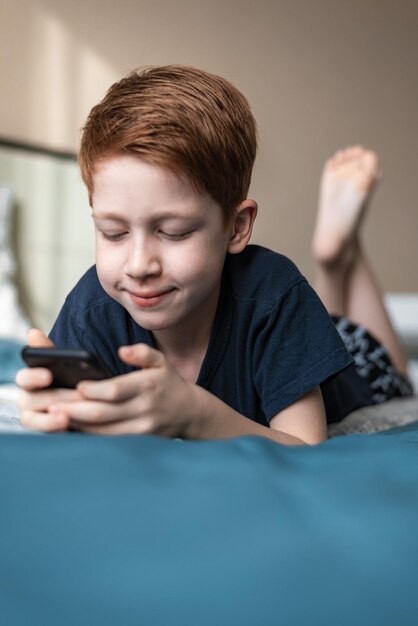 Menino ruivo criança segura um telefone celular nas mãos assiste desenhos animados e videogames Retrato de estilo de vida deita na cama em casa e ri fotografia vertical