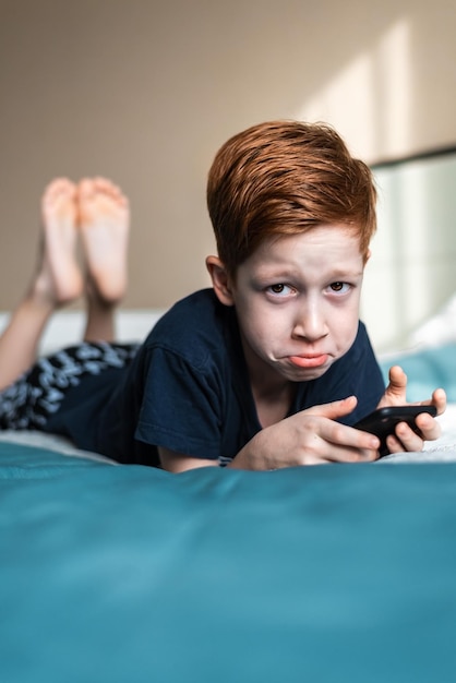 Menino ruivo criança segurando um telefone celular nas mãos chateado o retrato do estilo de vida está na cama em casa