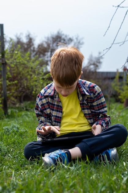 Menino ruivo feliz jogando no tablet ou assistindo desenhos na grama verde no quintal da aldeia