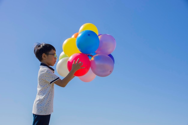 Foto menino segurando balões multicoloridos enquanto está de pé contra o céu azul claro