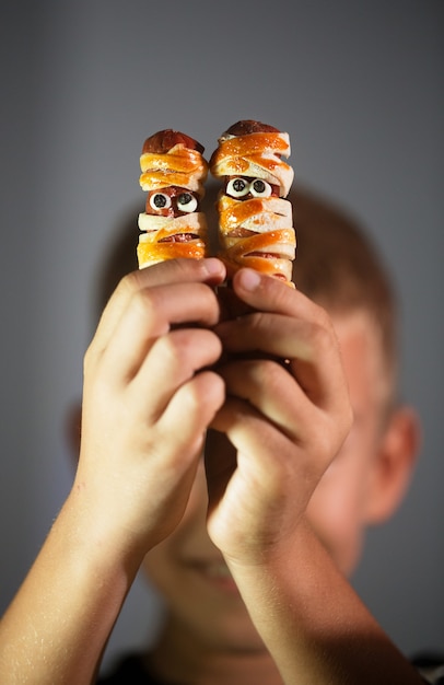 Foto menino segurando salgadinhos de halloween em formato de múmia, feitos de massa e salsicha