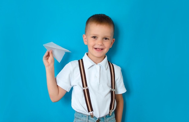 Menino segurando um avião de papel em um fundo azul Uma foto de um jovem estudante animado com um sorriso feliz e positivo joga um modelo de avião no ar