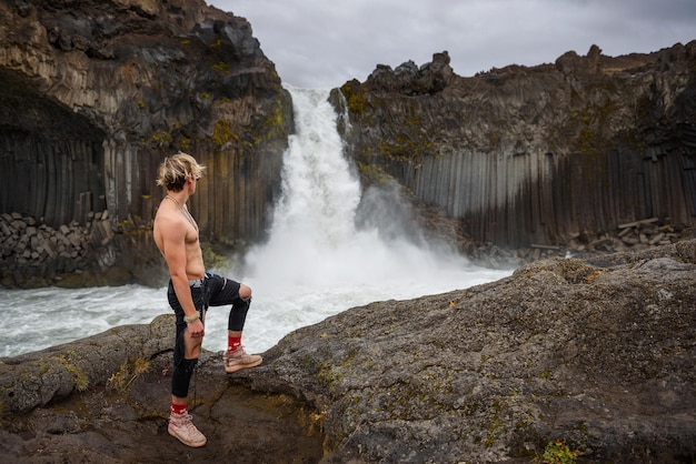 Menino sem camisa e musculoso olha para a cachoeira aldeyjarfoss na islândia